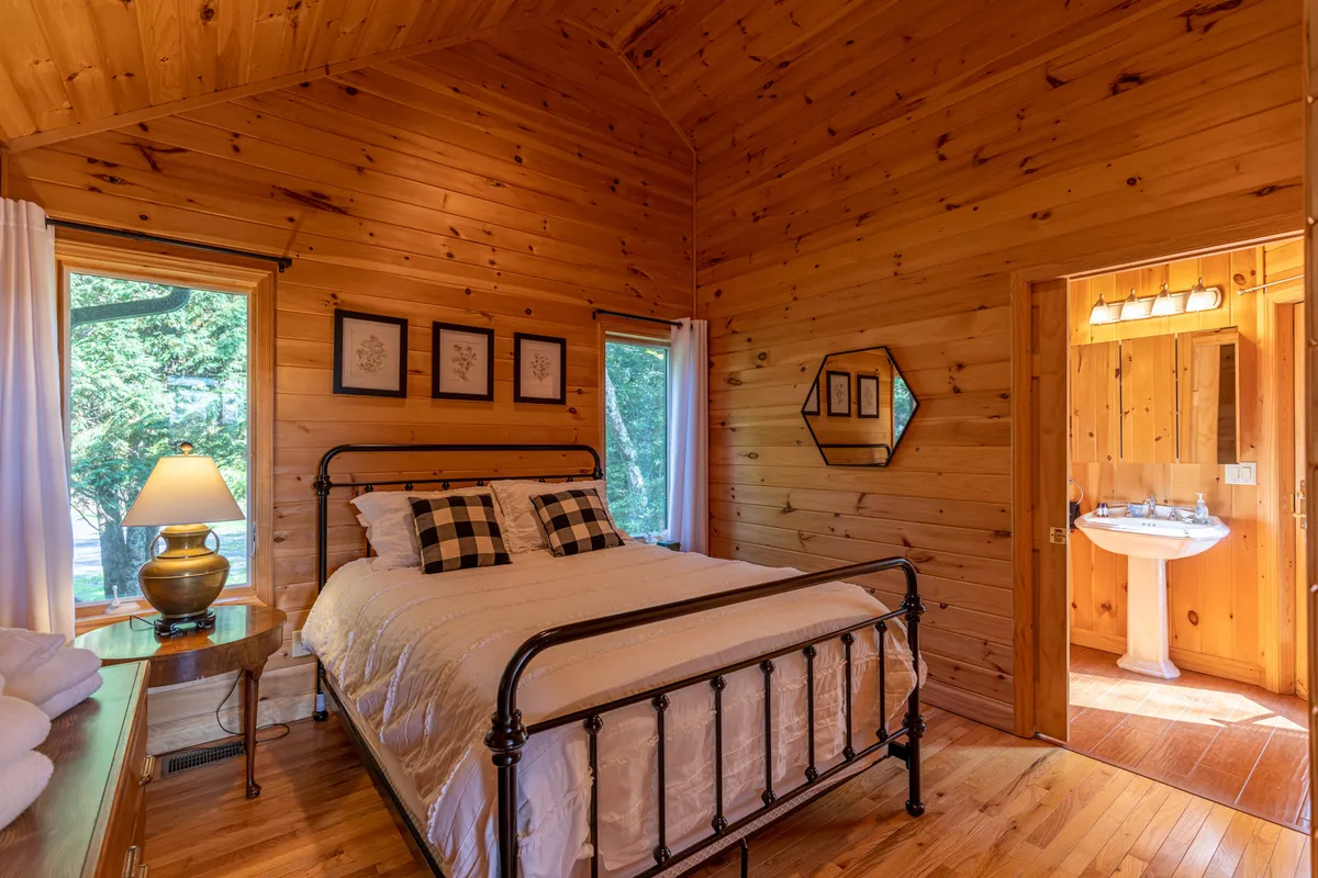 A cozy bedroom with a queen bed dressed in black and white checkered pillows sits against warm wooden walls. Large windows fill the room with natural light, while decorative wall art adds personality. The door to the bathroom is visible, offering a seamless connection between the sleeping area and bath.