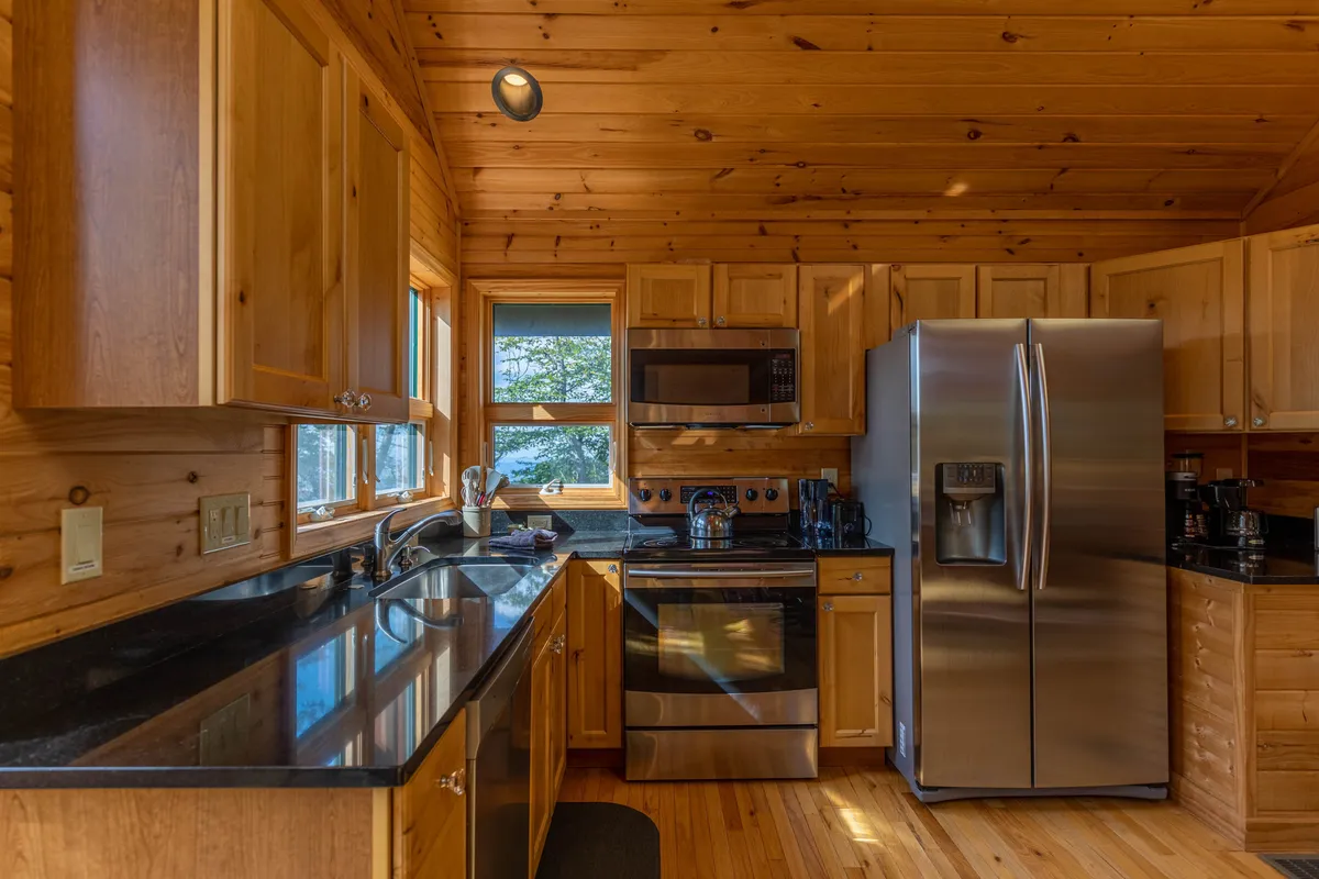 A view into the modern kitchen, this image centers on sleek stainless steel appliances, a black granite countertop, and warm wood cabinetry. A window above the counter lets in light and offers a scenic view, reinforcing the kitchen’s inviting, functional vibe. The space is staged for practical meal prep and highlights the complete cookware and utensils available.