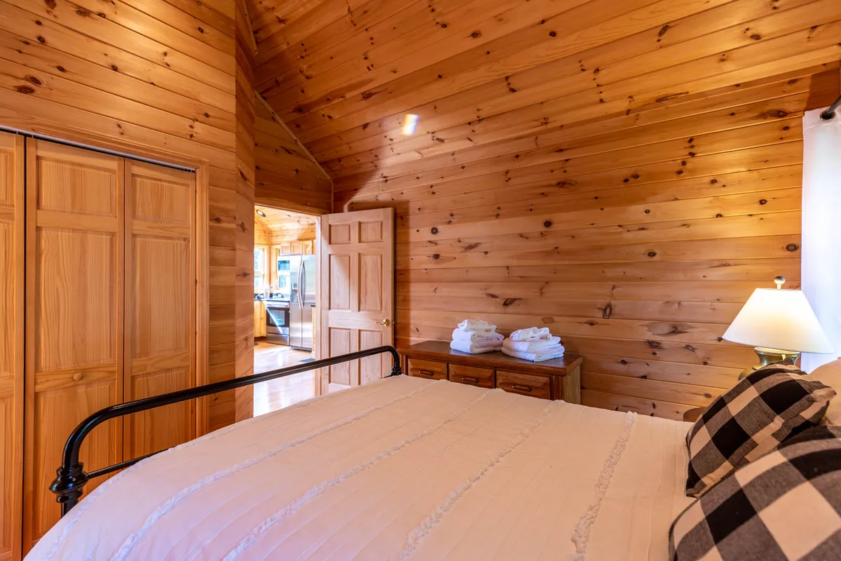 A serene bedroom scene featuring a queen bed with crisp white linens and checkered accent pillows. Wooden walls and furniture enhance the intimate, cabin-like atmosphere. Rolled towels rest atop a dresser, emphasizing comfort and readiness for relaxation.