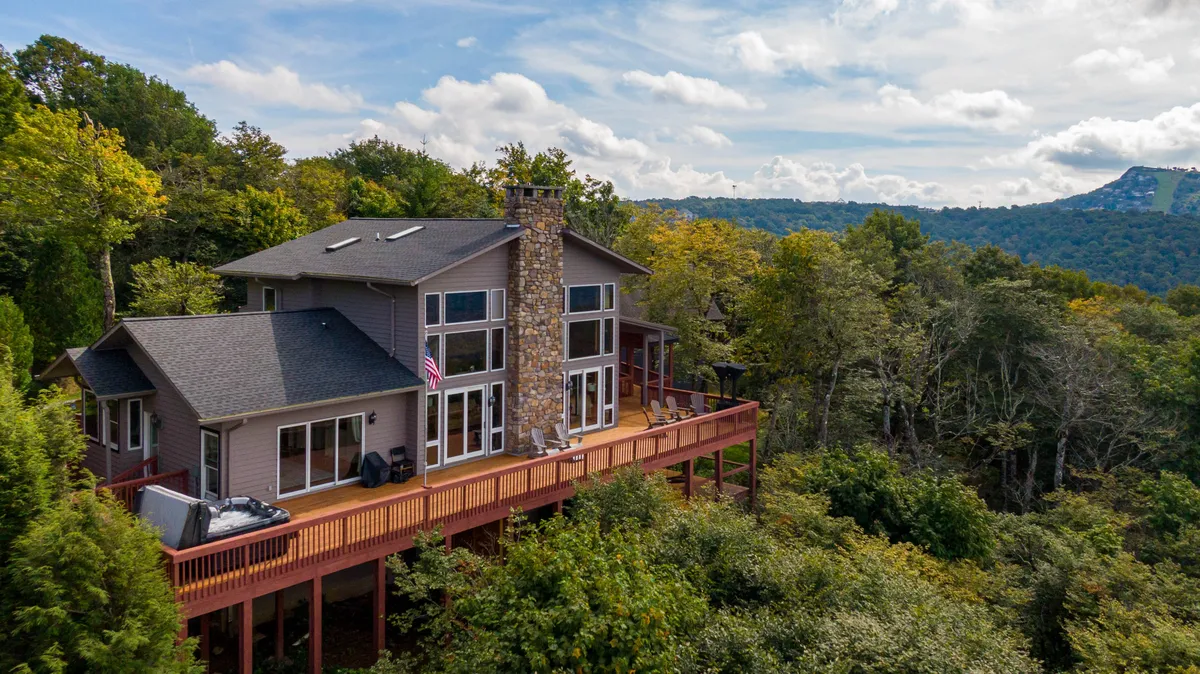 A wide exterior view shows the entire house perched above the garage, featuring a spacious wooden deck and a dramatic stone chimney. The backdrop includes dense trees and distant mountain peaks, situating the Chalet in its striking natural environment.
