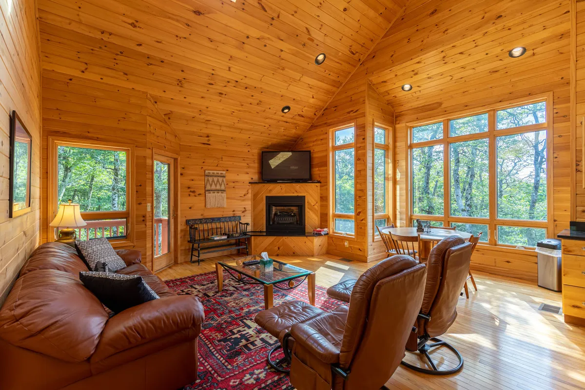 This image captures a welcoming living room with a prominent gas fireplace, a leather sofa, large windows streaming in natural light, a warm wooden ceiling, and a decorative rug. The arrangement and textures exude cozy mountain ambiance, with expansive views framed by the windows.