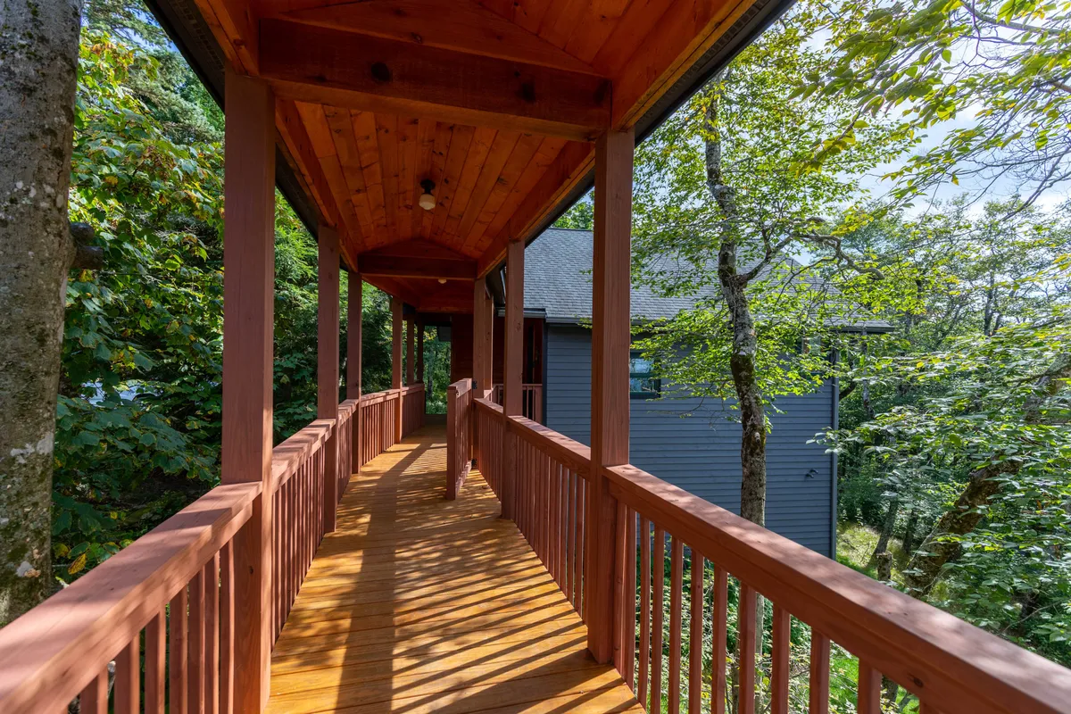 A rustic wooden walkway leads through lush greenery toward the house, with the roofline visible above the foliage. The scene captures the unique, elevated approach to the property and evokes a tranquil mountain setting. The walkway appears slightly elevated, suggesting the presence of architectural features like a skybridge.