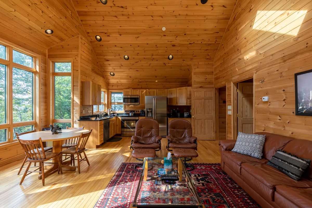 A cozy, open-concept living area featuring large windows with scenic nature views, leather armchairs arranged for intimate conversation or relaxation, a colorful area rug, a wooden dining table, and hints of the kitchen in the background. The room glows with the warmth of natural light and inviting decor.