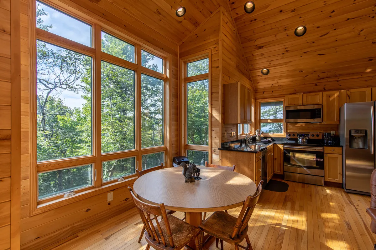 This image showcases a cozy, light-filled dining nook featuring a wooden table set beside expansive windows. The scene is oriented to highlight both the well-equipped kitchen—complete with modern appliances—and the inviting meal space, with natural light streaming in and framing a tranquil, mountain-inspired backdrop. The table setting hints at shared family meals or peaceful morning coffees.