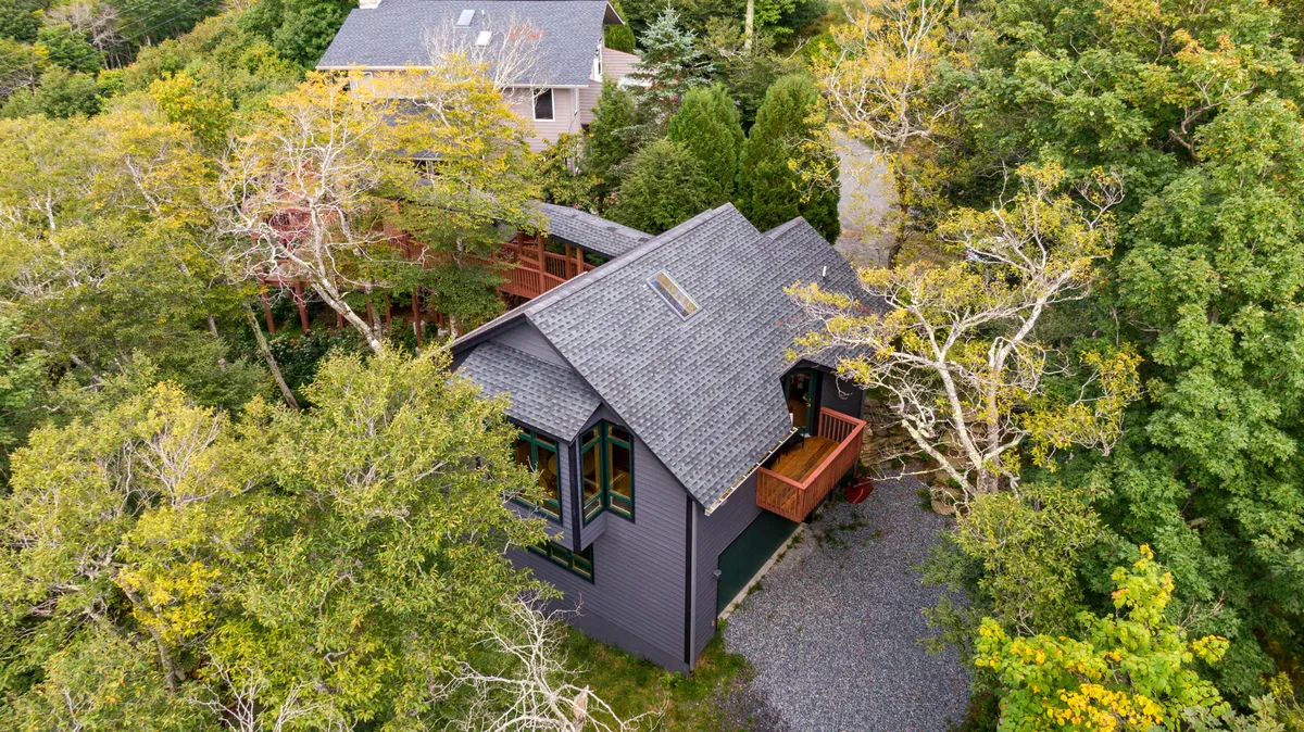 The image shows the distinctive chalet-style house nestled among tall trees, with a spacious deck offering panoramic sunset views. The serene mountain setting is emphasized by the lush greenery, and the inviting outdoor area hints at relaxing mornings with coffee or evenings enjoying nature.